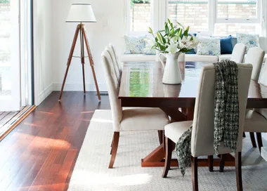 Dining room with a wooden table, cream chairs, floral centerpiece, tripod lamp, and window seat with cushions.