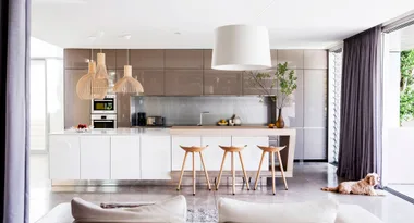 Modern kitchen with beige cabinets, white island, wooden stools, pendant lights, and a dog by the window.