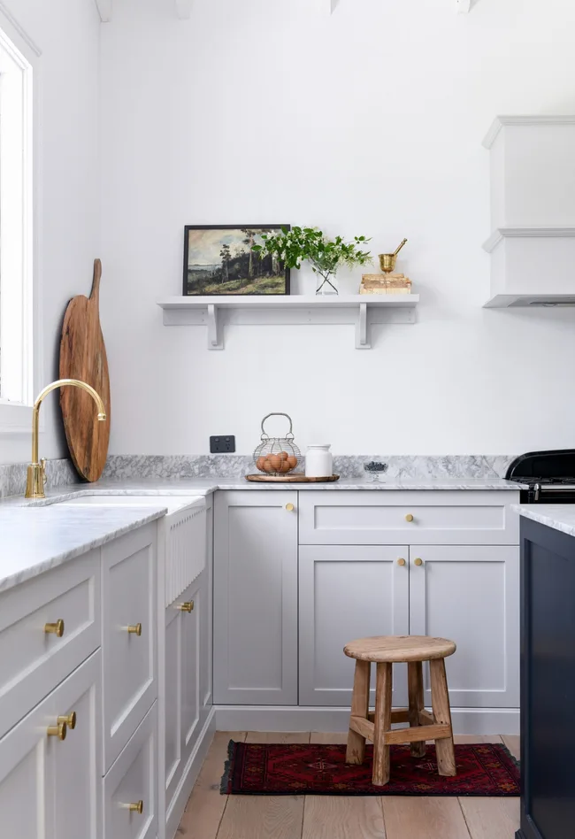 White kitchen with marble countertop, brass fixtures, wooden stool, cutting board, potted plant, and framed artwork.
