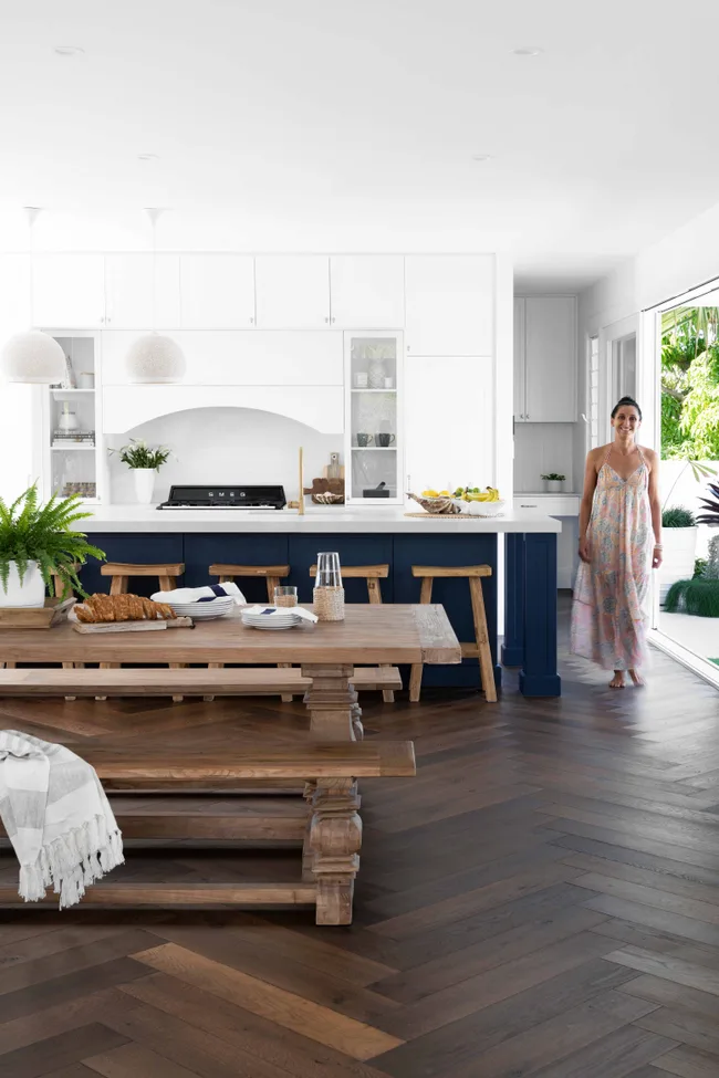 Bright kitchen with a wooden table, stools, plants, and a woman in a dress standing near a large window.