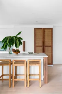 Modern kitchen with wooden stools, white countertop, leafy plant, and woven cabinet doors. Bright and minimalist design.