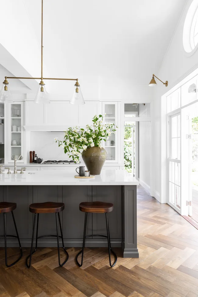 Bright kitchen with white cabinets, wooden stools at island, pendant lights, and a vase with greenery on the counter.