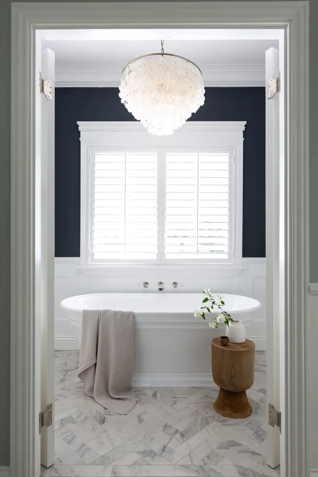Elegant bathroom with a freestanding tub, chandelier, wood stool, and flowers, framed by open white doors.