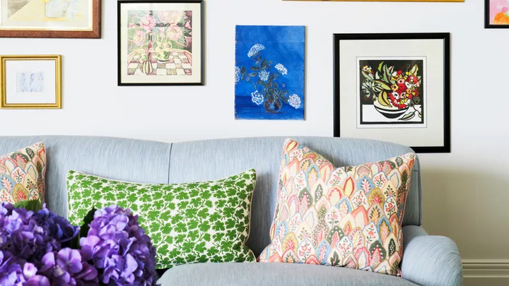 Colorful gallery wall above a light blue sofa with patterned pillows; vase of purple flowers on table in foreground.