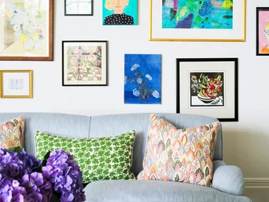 Colorful gallery wall above a light blue sofa with patterned pillows; vase of purple flowers on table in foreground.