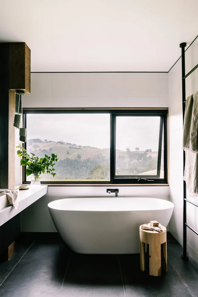 Modern bathroom with freestanding tub, large window view of rolling hills, and wooden stool.