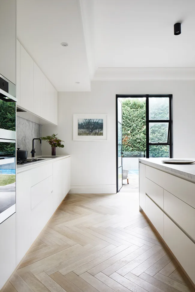 Modern kitchen with white cabinets, herringbone wood floor, and open door to garden.