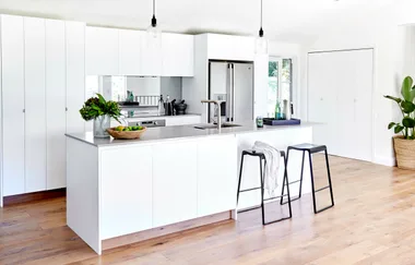Modern white kitchen with island, two bar stools, pendant lights, and wooden floor. Green plant decor on the counter.