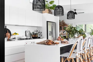 Modern white kitchen with pendant lights, rattan chairs, plants, and bread on a large island countertop.