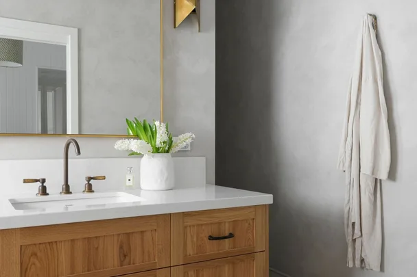Bathroom vanity with wooden cabinet, white countertop, green flowers, brass faucet, and hanging towel on gray wall.