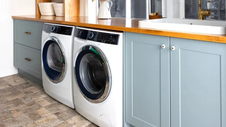 Laundry room with a washing machine and dryer under a wooden countertop, adjacent to a sink and light blue cabinets.