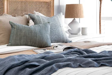 Cozy bedroom with a striped bedspread, blue and beige pillows, a magazine, and a lamp on a white side table.