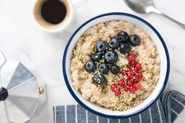 Bowl of oatmeal topped with blueberries, red currants, and seeds, next to a cup of coffee and a coffee maker.
