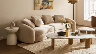 A beige curved sofa with cushions, abstract art, and round tables with decor in a cozy, well-lit living room.