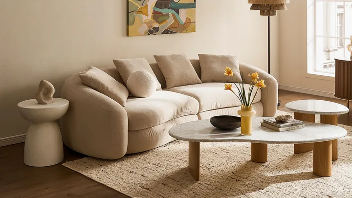 A beige curved sofa with cushions, abstract art, and round tables with decor in a cozy, well-lit living room.