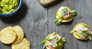 Tostadas topped with guacamole, sliced salmon, cucumber, red onion, and cilantro on a wooden surface.