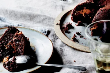 Slice of chocolate cake with caramel filling on a plate, next to a larger cake portion on a platter.