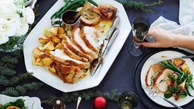 A festive meal with sliced roasted turkey, potatoes, and greens on a plate; hand holding a glass, flowers nearby.