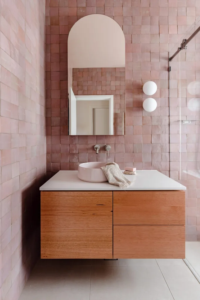 A main bathroom featuring pink zellige wall tiles, white floor tiles, wooden joinery, a round basin, an arched mirror and two round wall sconces beside a shower. The tapware is brushed nickel in a curved style.