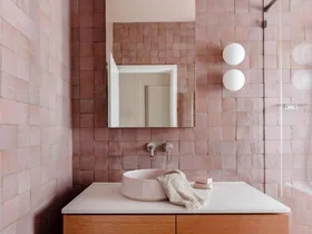 A main bathroom featuring pink zellige wall tiles, white floor tiles, wooden joinery, a round basin, an arched mirror and two round wall sconces beside a shower. The tapware is brushed nickel in a curved style. The bathroom design is cohesive.