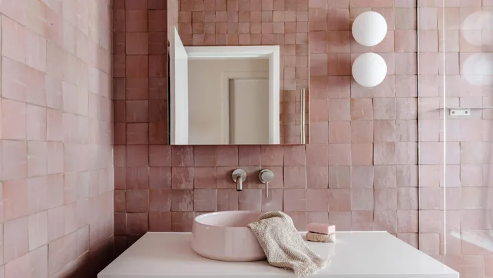 A main bathroom featuring pink zellige wall tiles, white floor tiles, wooden joinery, a round basin, an arched mirror and two round wall sconces beside a shower. The tapware is brushed nickel in a curved style. The bathroom design is cohesive.