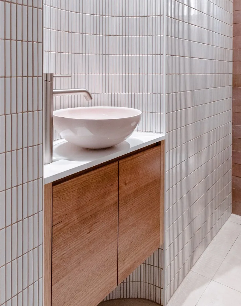 A powder room vanity, set into a curved inlet. It features pink zellige wall tiles, white kit kat wall tiles, white floor tiles, wooden joinery, a round basin and tapware in brushed nickel in a curved style.