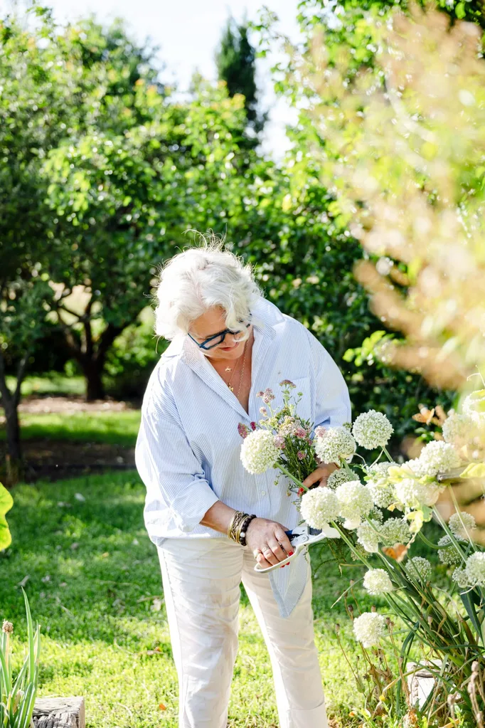 A woman wears white pants and a light blue buttoned shirt while using secateurs to cut flowers from her garden.