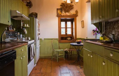 Cozy kitchen with green cabinets, wooden chairs, rustic window, and a small round table with a fruit bowl.
