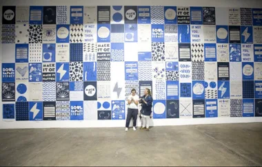 Two people stand in front of a large blue and white mural with motivational phrases and geometric patterns.