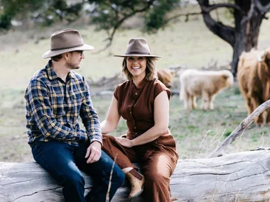 Edwina Bartholomew and Neil Varcoe on a log with pet cows in the background.