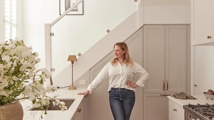 Woman in a white shirt leans against a kitchen counter with flowers in the foreground and a staircase behind her.