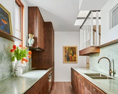 A galley kitchen with a light green quartzite that is used as the benchtop and the splashback, with underbench joinery made from hand-stained dark timber oak veneer. The tapware is also dark, in Antique Brass, with wooden flooring and framed paintings on the wall and an open shelf.