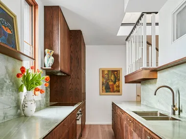 A galley kitchen with a light green quartzite that is used as the benchtop and the splashback, with underbench joinery made from hand-stained dark timber oak veneer. The tapware is also dark, in Antique Brass, with wooden flooring and framed paintings on the wall and an open shelf.