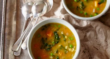 Bowls of vegetable soup with peas, greens, and carrots on a cloth, accompanied by silver spoons on a tray.