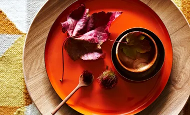 A wooden bowl and spoon on an orange plate with leaves and a small fruit, set on a wooden surface.
