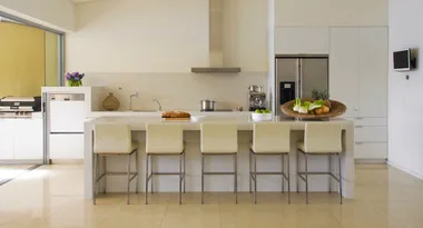 Modern kitchen with white cabinets, island with five beige stools, basket of fruits, and stainless steel appliances.