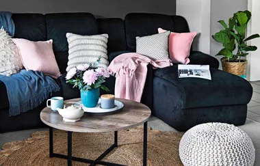 Cozy living room with a black couch, colorful cushions, a round table with tea set and flowers, and a potted plant.