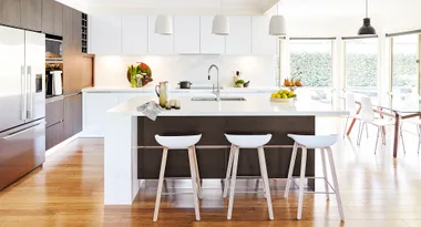 Modern kitchen with wooden floors, large island, white stools, pendant lights, and a view of greenery through windows.