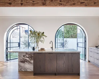 A kitchen with oak timber floorboards, oak veneer joinery, structural timber beams along the white ceiling, marble benchtops, marble splashbacks and two arched French doors in glass and black steel. The kitchen is open plan with a sizable island in the middle.