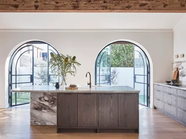 A kitchen with oak timber floorboards, oak veneer joinery, structural timber beams along the white ceiling, marble benchtops, marble splashbacks and two arched French doors in glass and black steel. The kitchen is open plan with a sizable island in the middle.