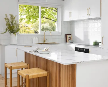 A white kitchen with timber details, including a ribbed island profile and woven bar stools.