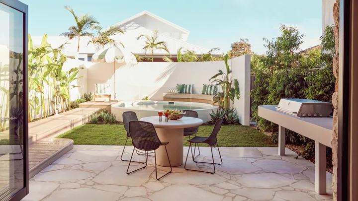 An alfresco dining area with a barbecue with a curved white wall around a circular plunge pool in the background. Green and white striped cushions decorate the bench around the pool and stone is used for the flooring of the outdoor dining area.