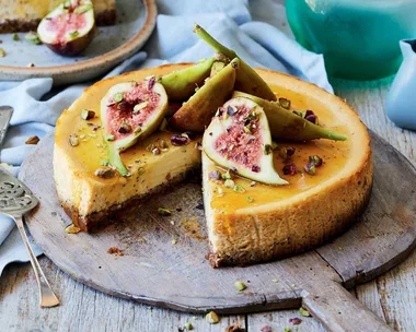 Cheesecake topped with fresh figs and chopped pistachios on a wooden board, slice removed, ornate silver serving utensils beside.