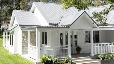White house with a metal roof, front porch, green lawn, and potted plants, set in a tree-lined area.