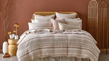 Cozy bedroom with a striped bedspread, multiple pillows, wooden side table, vase with flowers, and a rattan room divider.