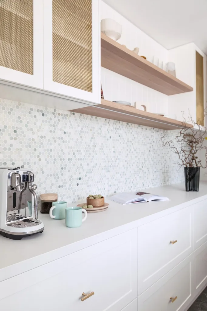 A mosaic tiled splashback and a white benchtop, with white underbench cabinetry and upper cabinetry that has brass mesh profiles. There is open shelving made of timber.