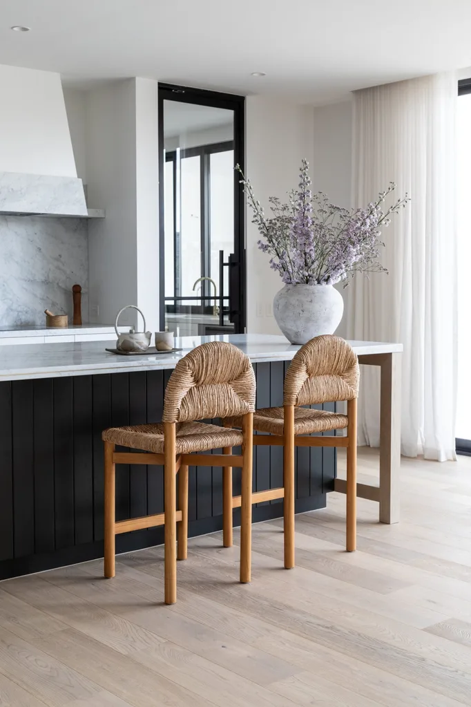 A sizable kitchen island, topped with carrara marble and with sides that are finished in Porter's Paints Black Cockatoo. Bar stools are timber with woven detailing on the seat and cushioned back. The white rangehood cover has a strip of carrara marble around the base, with the same marble used as the splashback.