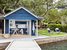 The blue cladded boatshed and two deck chairs on a lawn facing the bay.