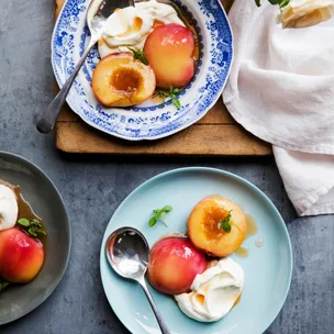 Peach halves with syrup and whipped cream on blue and gray plates, garnished with mint leaves, on a textured surface.