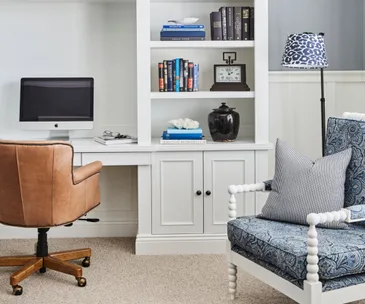 A home office with a computer, leather chair, bookshelves, decorative items, and a blue-patterned armchair.